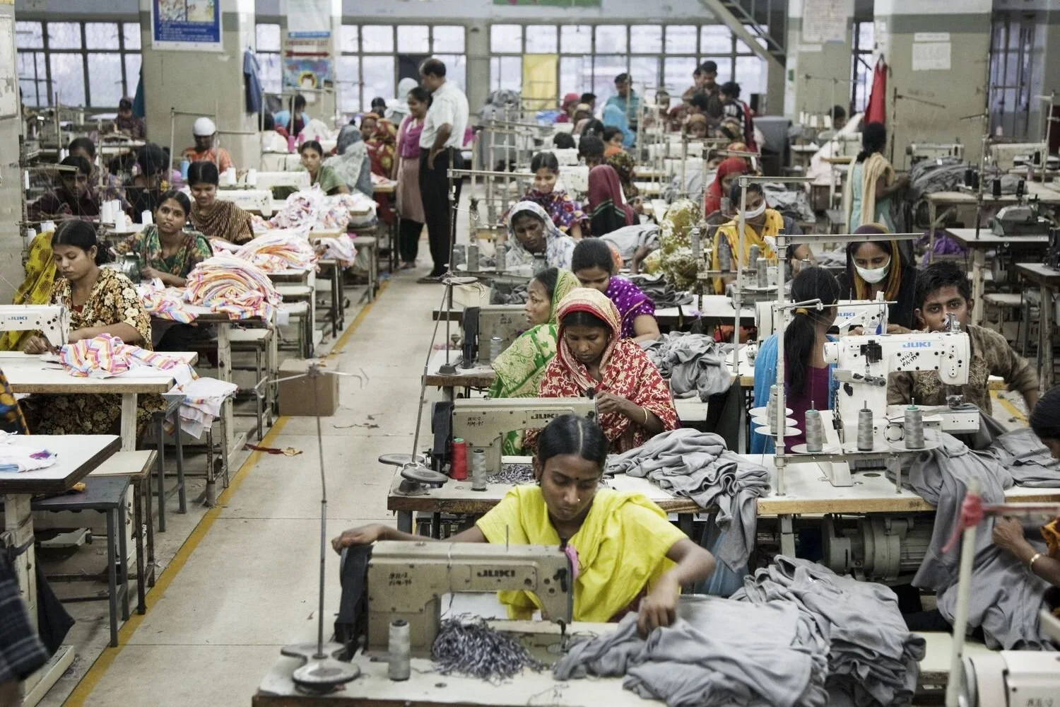 Un gran grupo de trabajadores de una fábrica de ropa están sentados frente a máquinas de coser en un taller abarrotado, cosiendo ropa y manipulando telas, muchos de ellos concentrados en sus tareas.